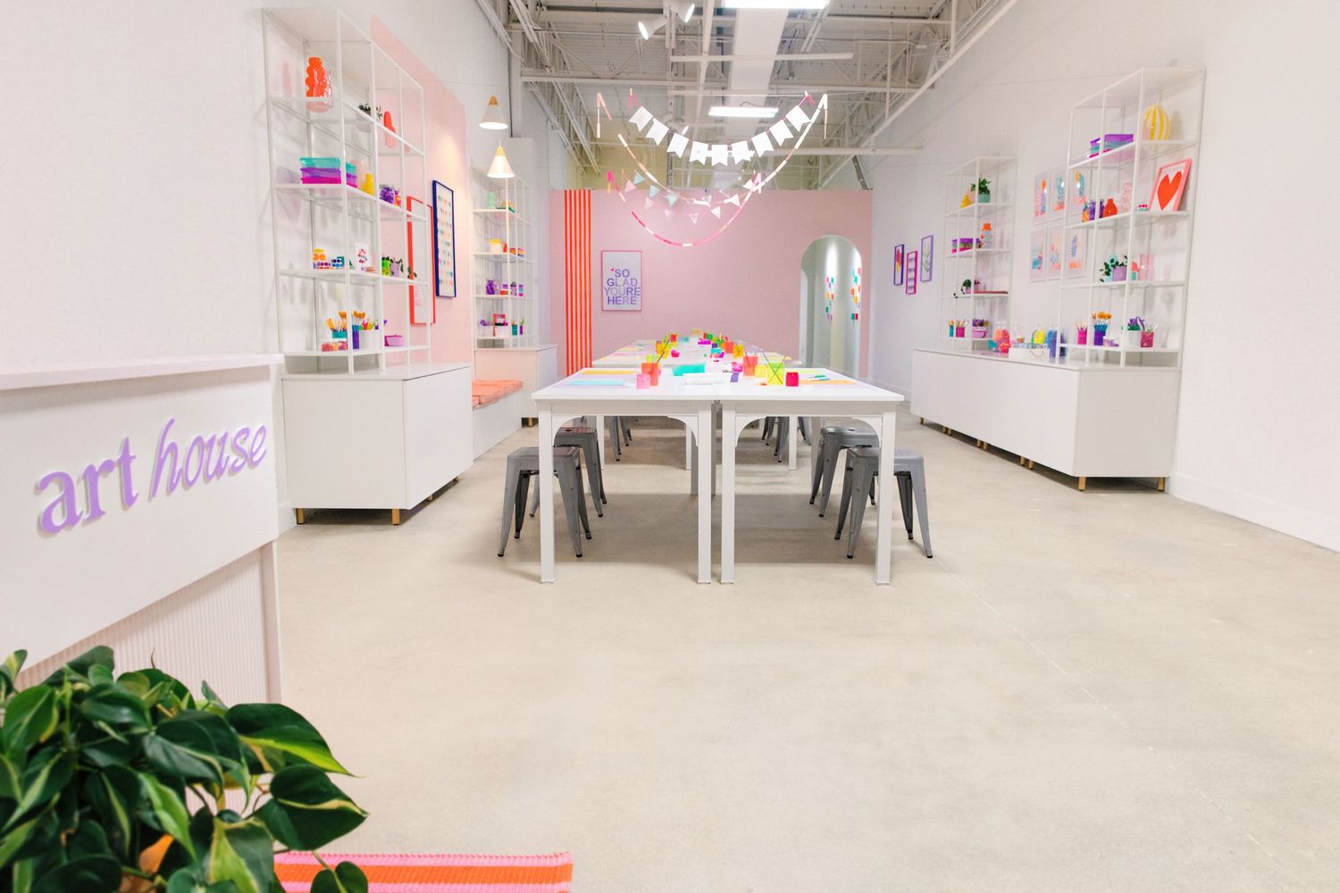 Bright art studio with white tables, colorful supplies on shelves, and pastel pink walls at Art House.