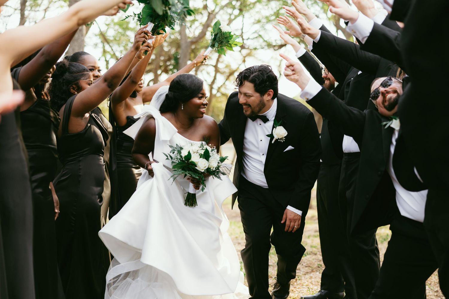 Bride and groom walk through a tunnel of raised hands holding greenery bouquets, surrounded by wedding party in black attire.