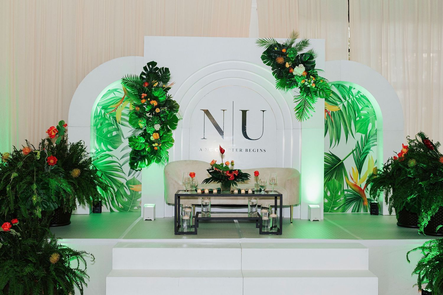 Elegant tropical wedding backdrop with white arch, lush greenery, monstera leaves, and floral arrangements framing an NU monogram sign.