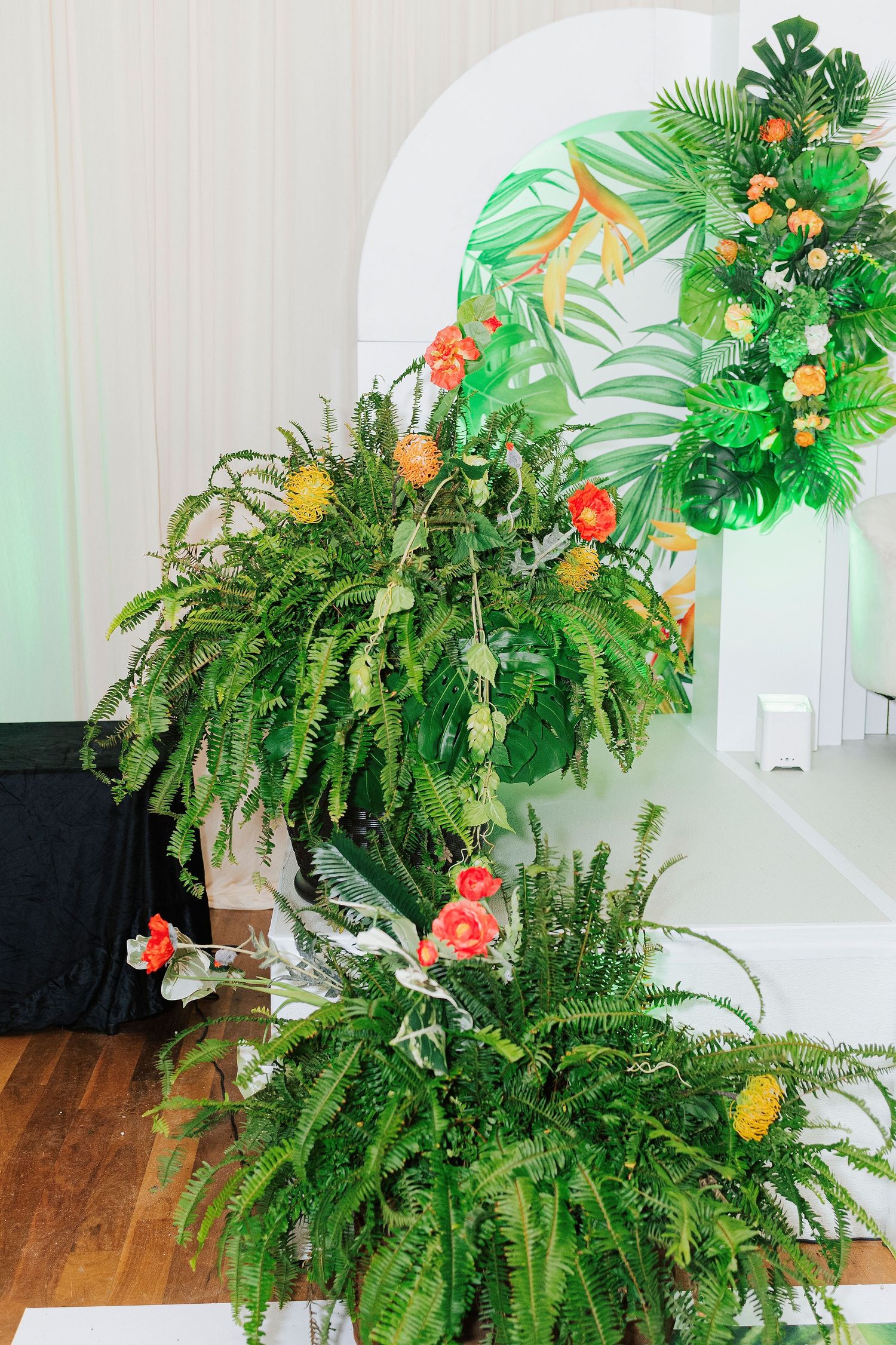 Tropical floral arrangement with ferns, orange flowers, and monstera leaves decorating a white arch backdrop.