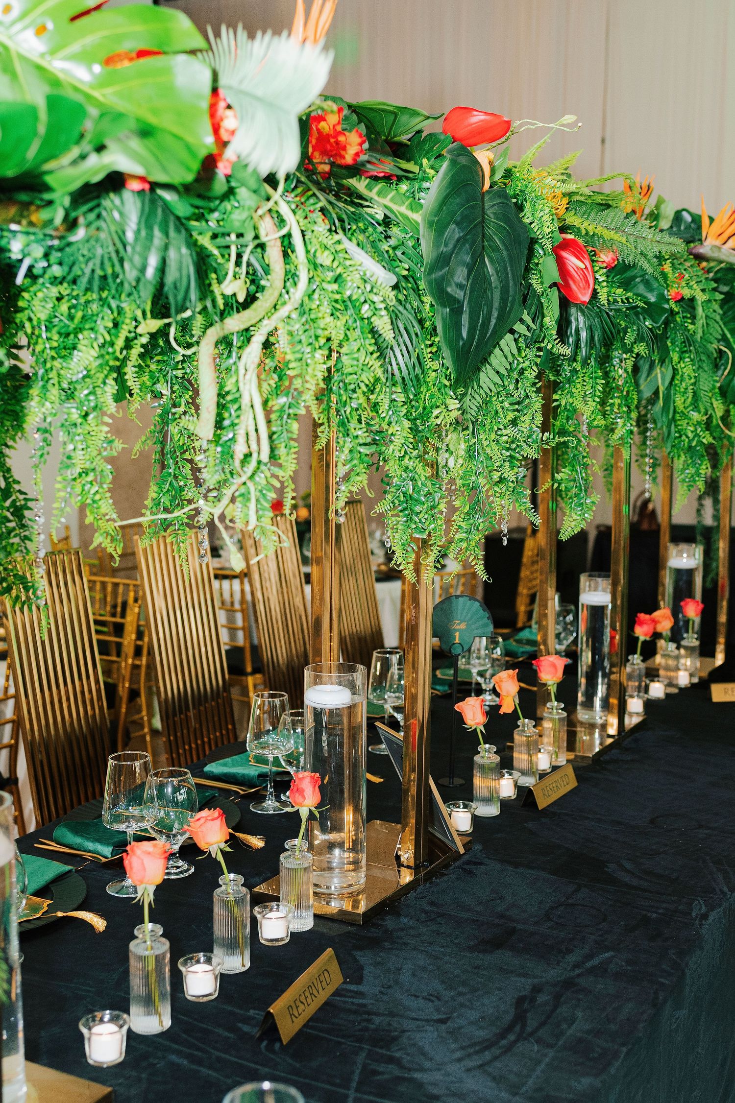 Tropical wedding reception table with lush greenery, gold stands, coral roses, and elegant glassware on black linen.