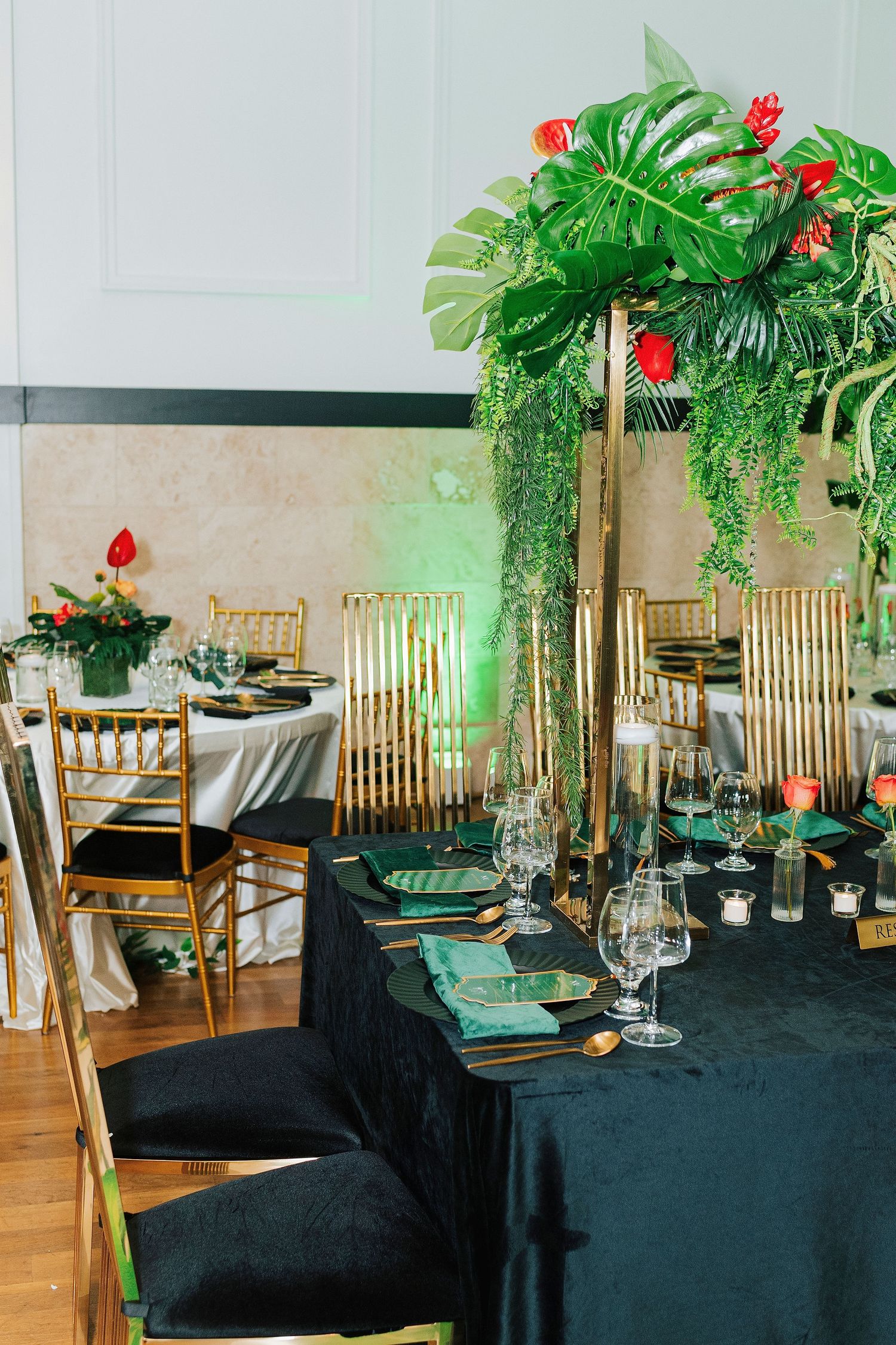 Elegant tropical wedding reception with black tablecloths, gold chiavari chairs, and tall green palm leaf centerpieces with red flowers.