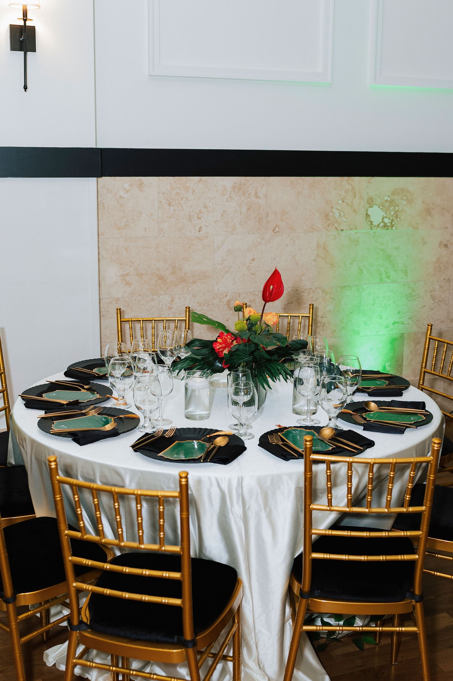 Elegant round banquet table with gold Chiavari chairs, black charger plates, crystal glassware, and tropical floral centerpiece.