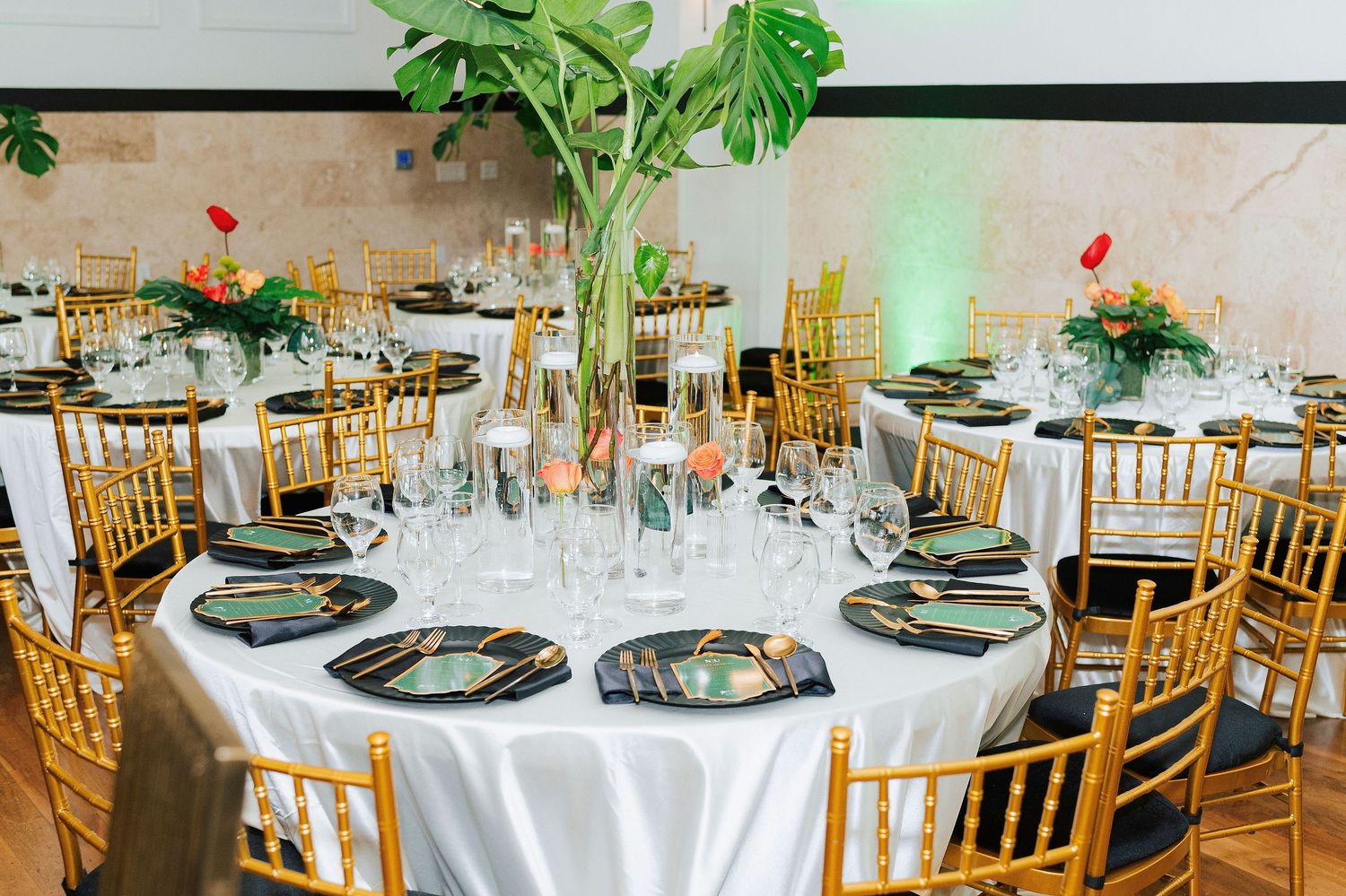 Elegant wedding reception with gold Chiavari chairs, white tablecloths, tropical leaf centerpieces, and crystal glassware.