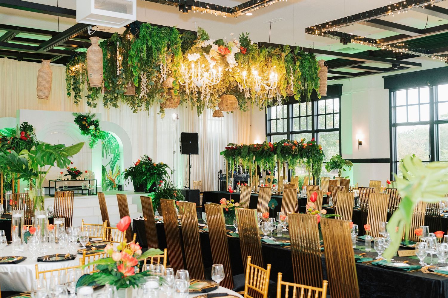 Elegant tropical wedding reception with lush green hanging foliage, gold chiavari chairs, and floral centerpieces in a bright venue.