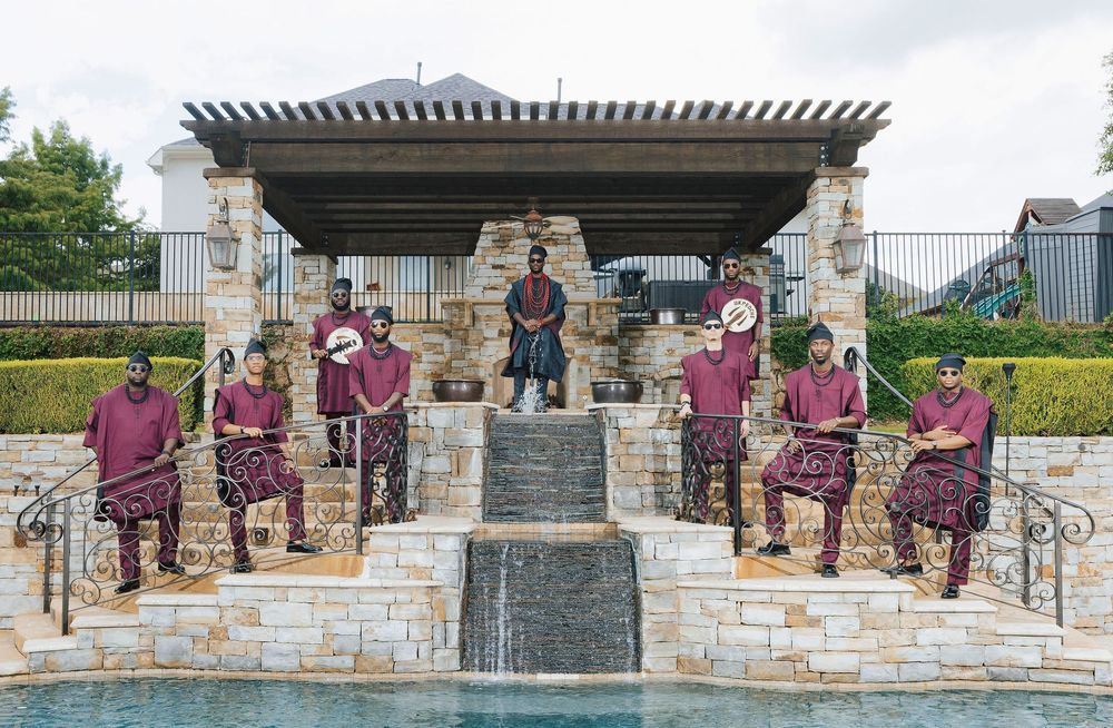 Marching band members in burgundy uniforms pose by a stone fountain with pergola backdrop.