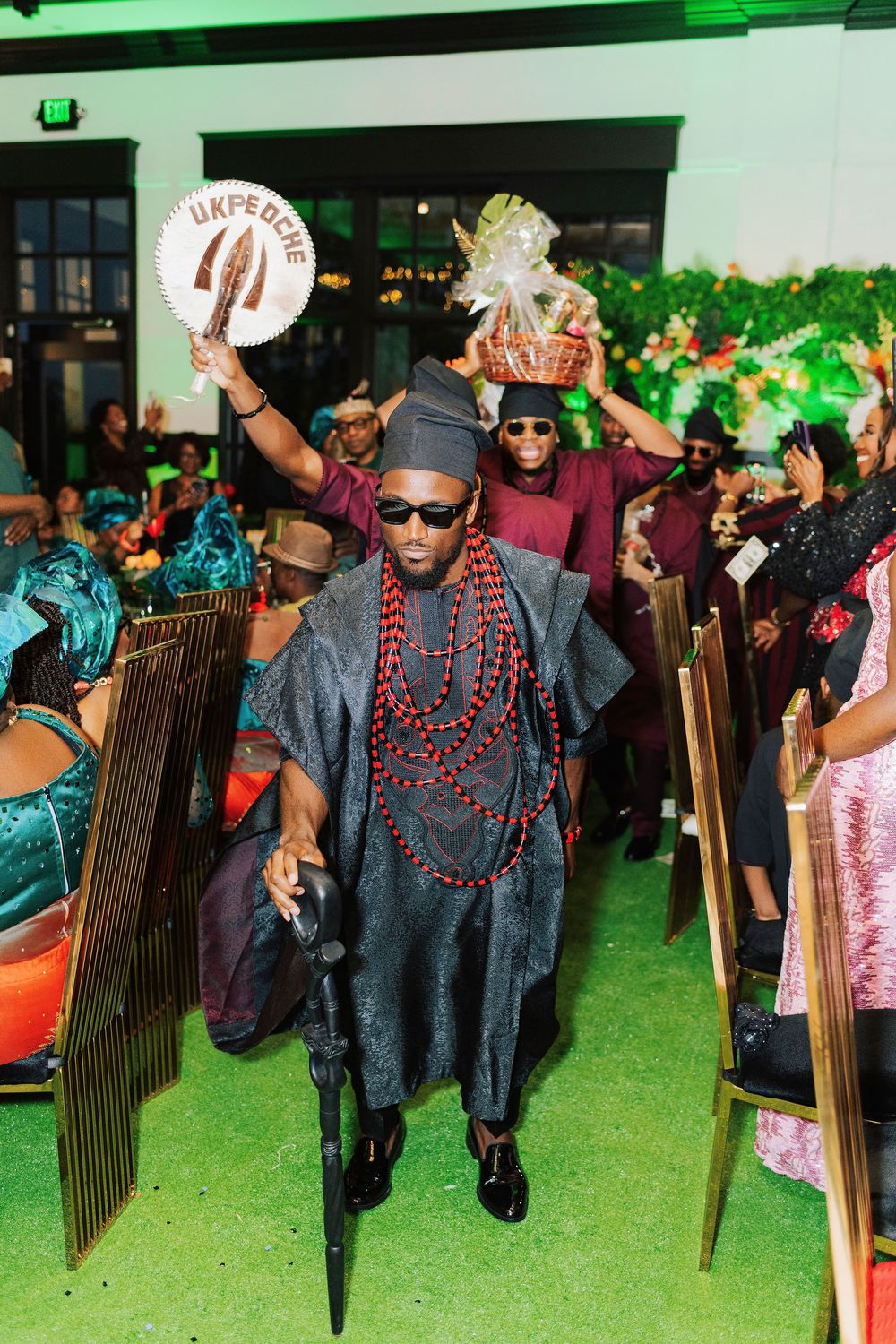 Man in traditional Nigerian attire with beads and cap leads a celebratory procession at a cultural wedding ceremony.