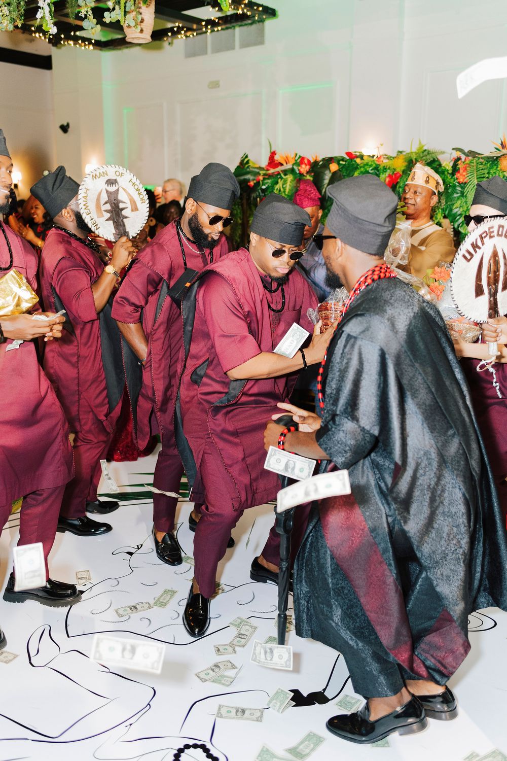 Men in burgundy traditional Nigerian attire dancing and spraying money at a vibrant cultural wedding celebration.