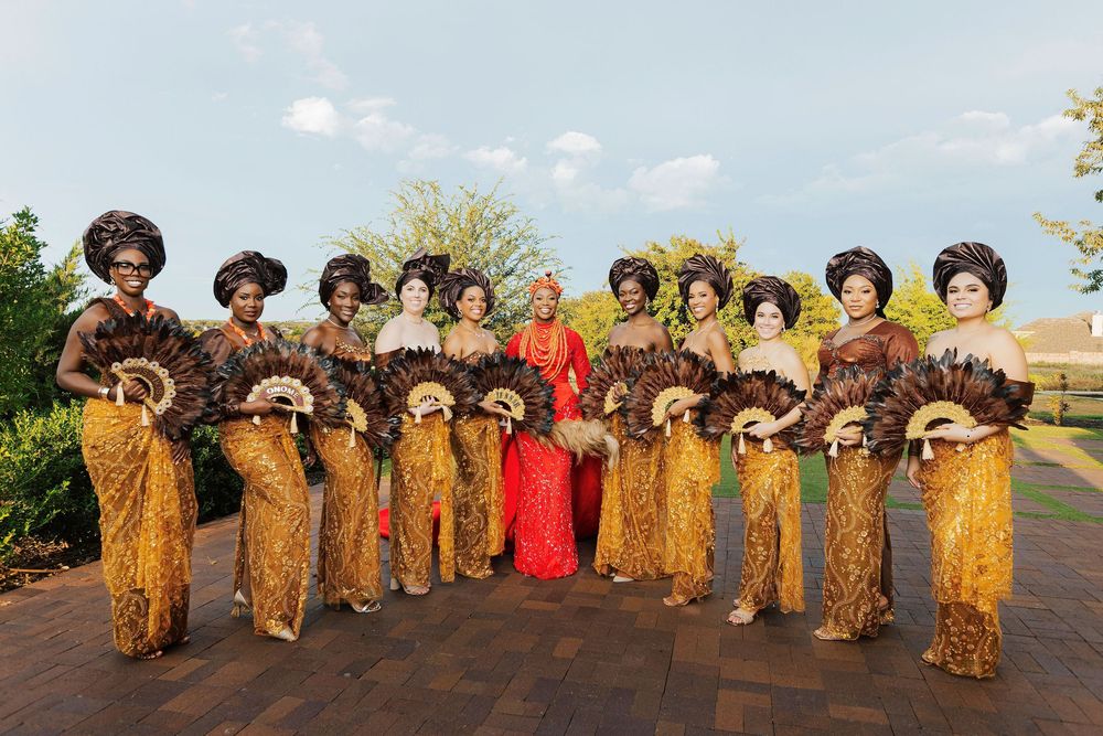 Bridal party in traditional Nigerian attire with gold and brown outfits, holding feather fans, posing outdoors.