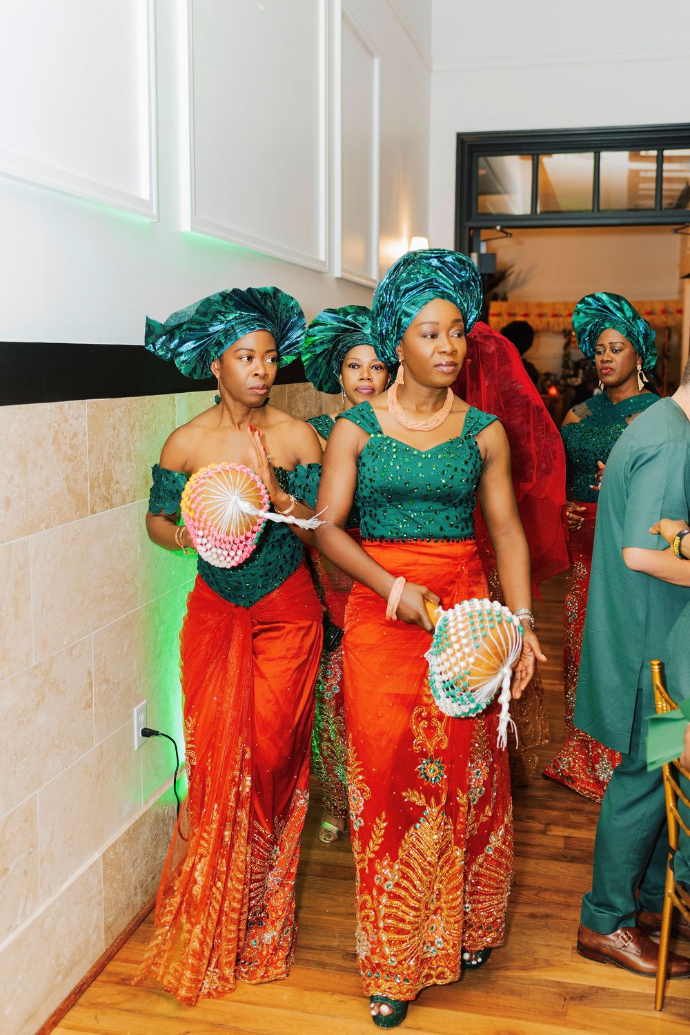 Women in traditional Nigerian attire with green headwraps and orange skirts walk in a cultural wedding ceremony.