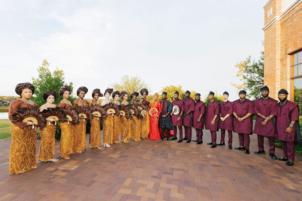 Large wedding party in traditional African attire, gold and maroon outfits, posing outdoors near brick building.