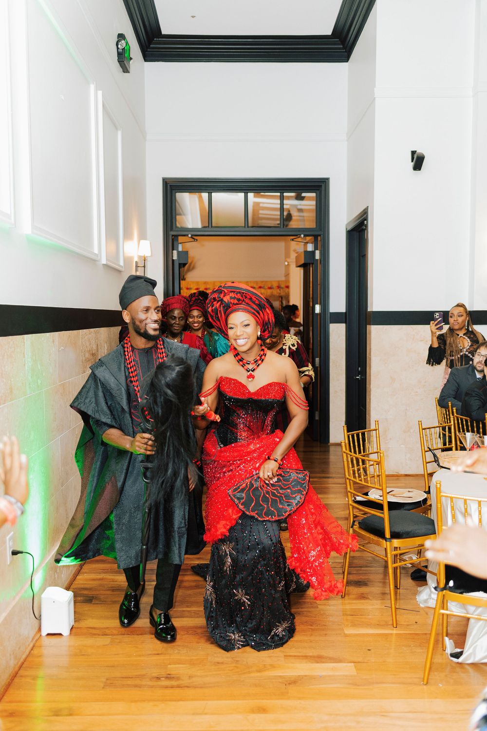 Couple in traditional Nigerian attire, red and black lace gown with gele headwrap, making grand entrance at wedding reception.