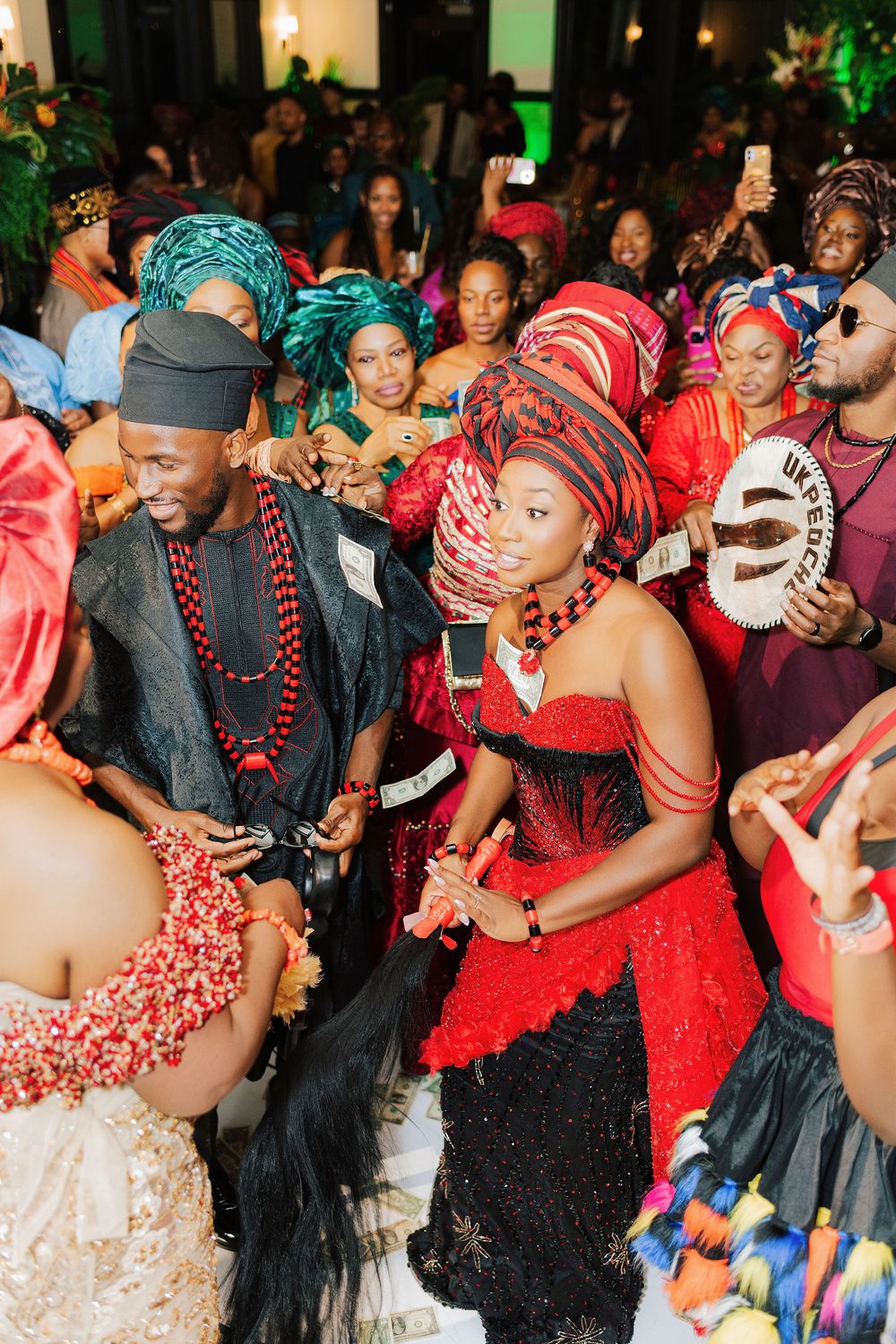 Vibrant Nigerian traditional wedding celebration with guests in colorful attire, bride in red beaded dress and gele headwrap.