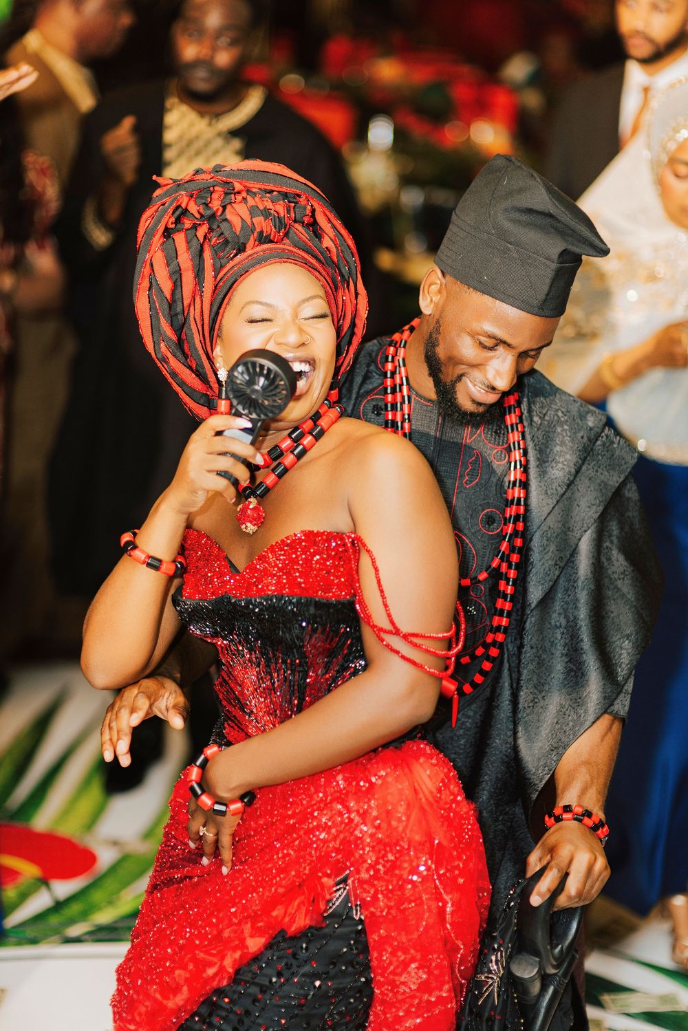 Couple in traditional Nigerian attire, woman in red sequin dress and gele headwrap singing at cultural wedding celebration.