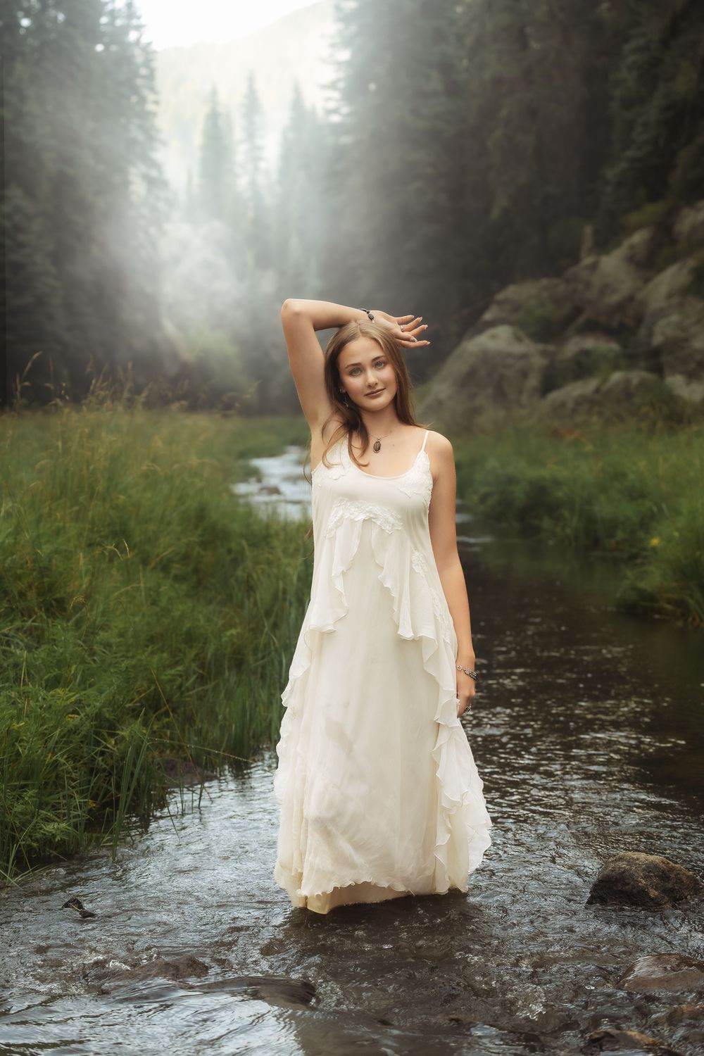Young woman in white ruffled dress standing in a misty forest stream surrounded by lush greenery.