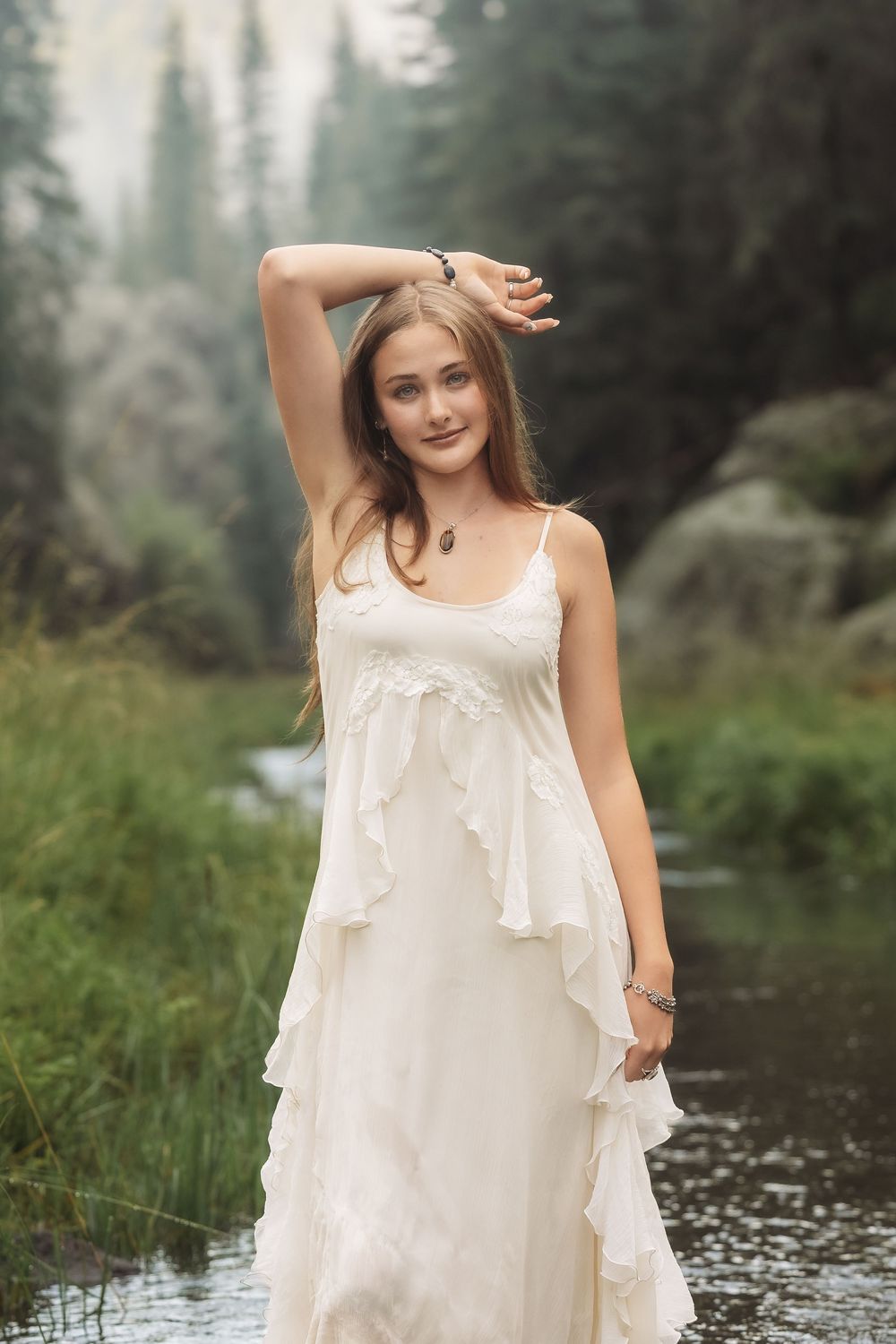 Young woman in white ruffled dress standing in forest stream with misty trees background.