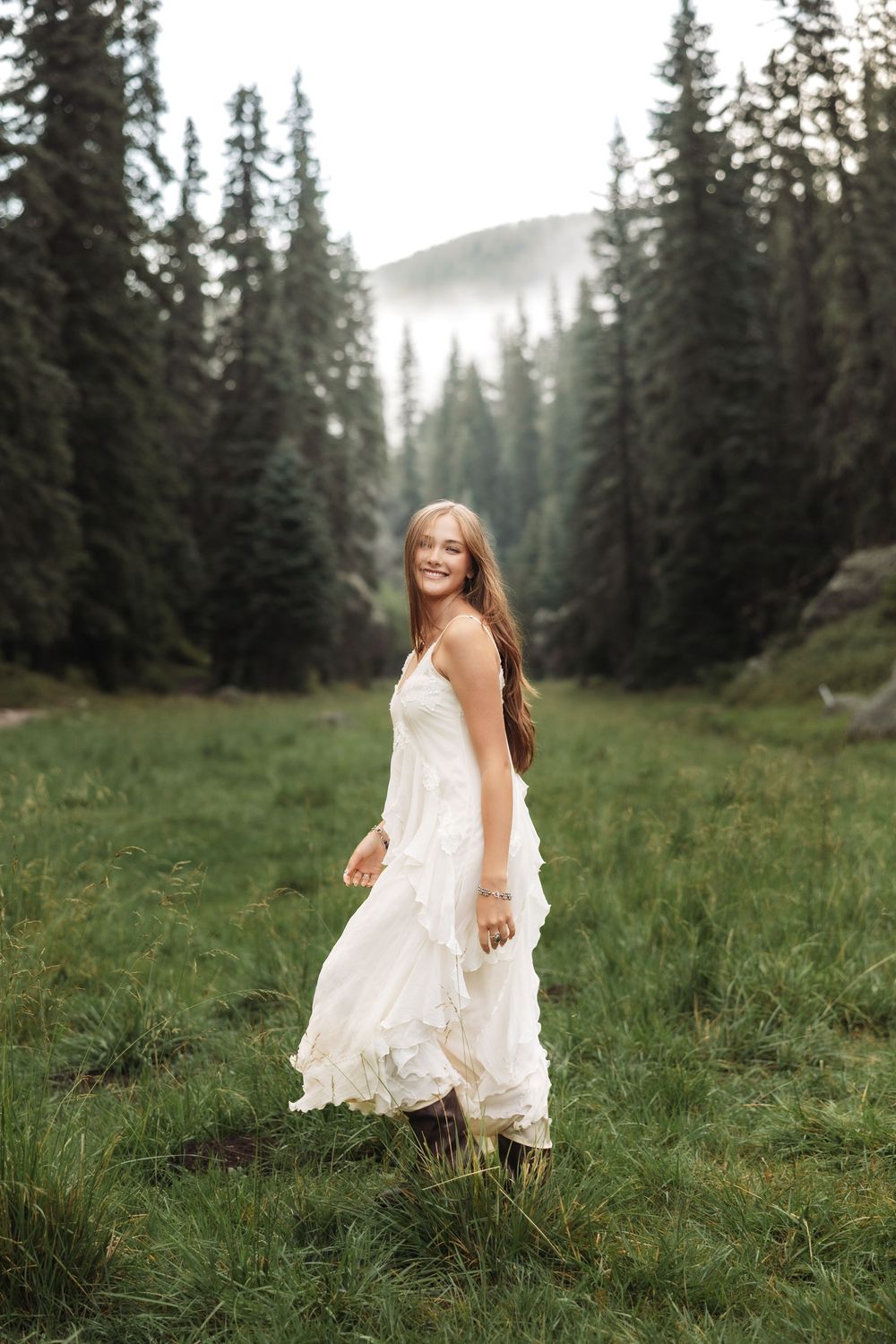 Young woman in white flowing dress smiling in a lush green mountain meadow surrounded by tall pine trees.
