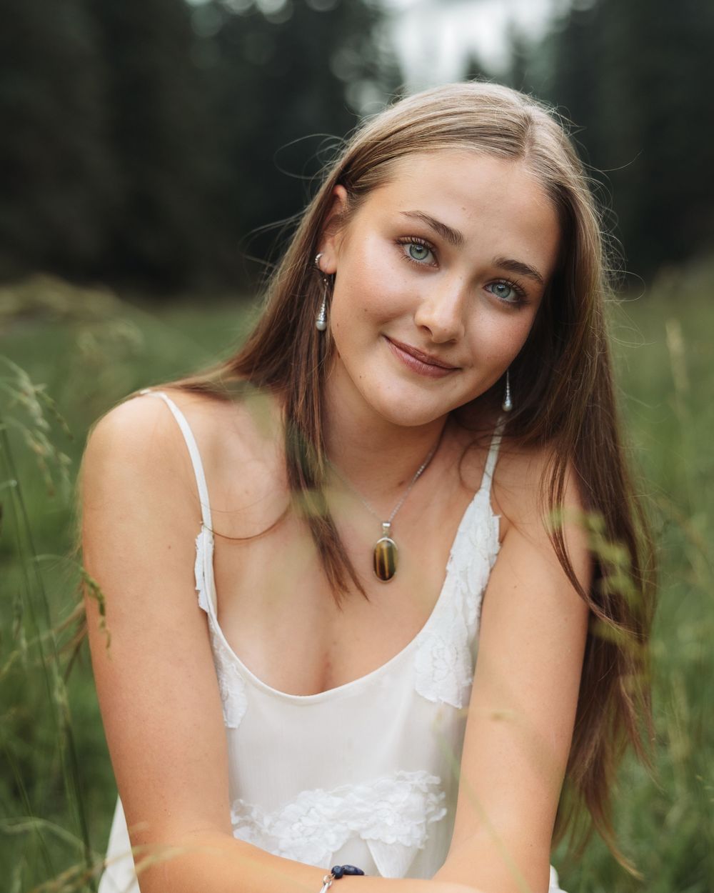 Young woman with long brown hair and blue eyes smiling outdoors in a field, wearing a white dress and pendant necklace.