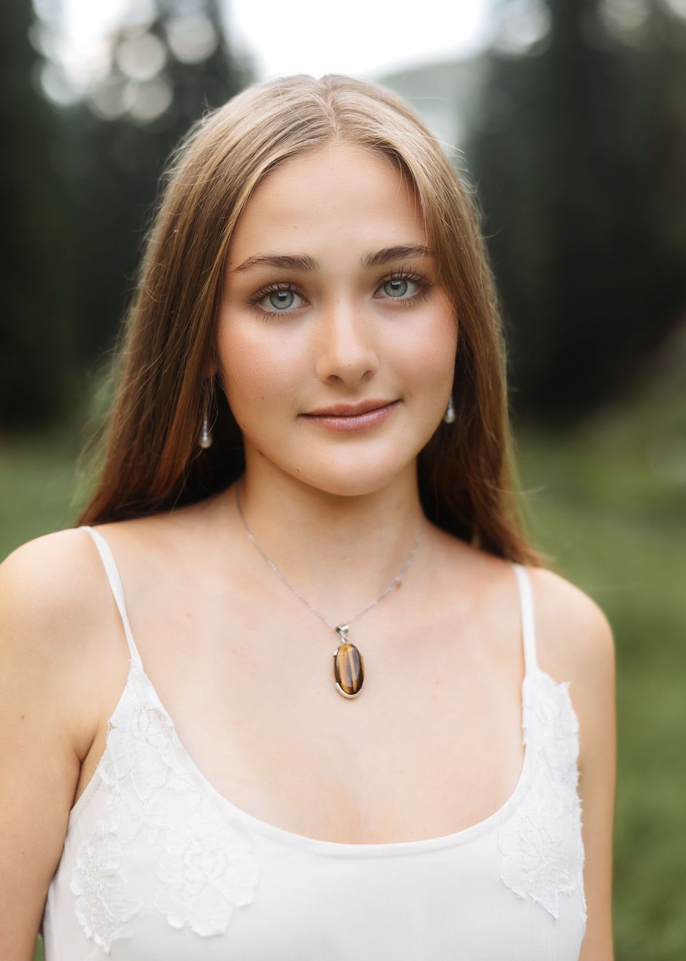 Young woman with long blonde hair and blue eyes wearing a white top and oval pendant necklace outdoors.