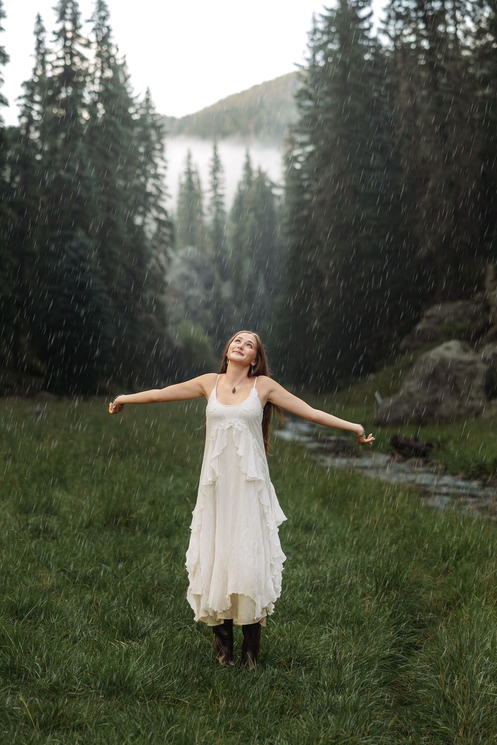 Woman in white dress standing in rain with arms outstretched, surrounded by pine forest and mountains.
