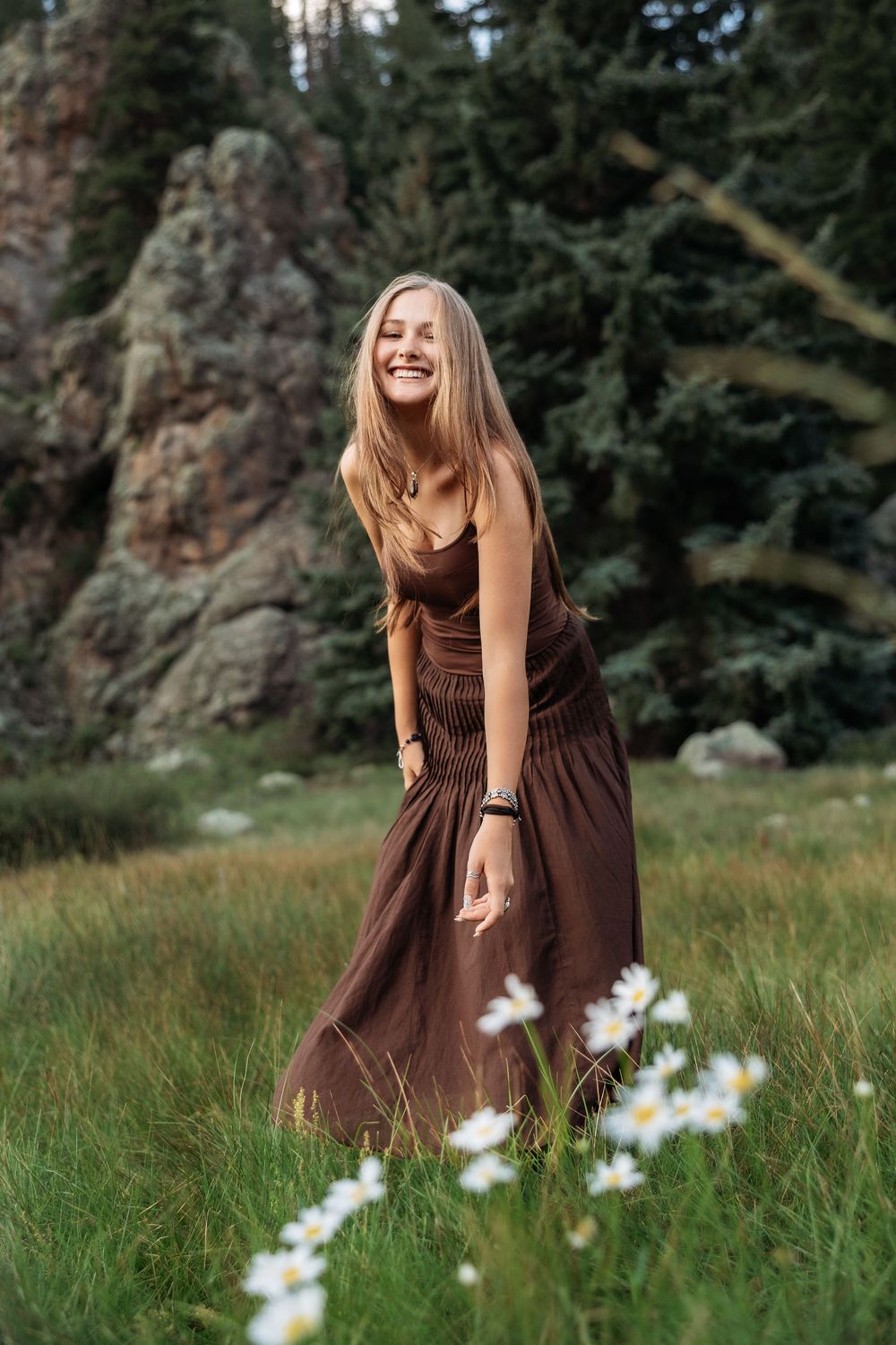 Smiling blonde woman in brown maxi dress standing in a meadow with daisies, rocks, and pine trees.