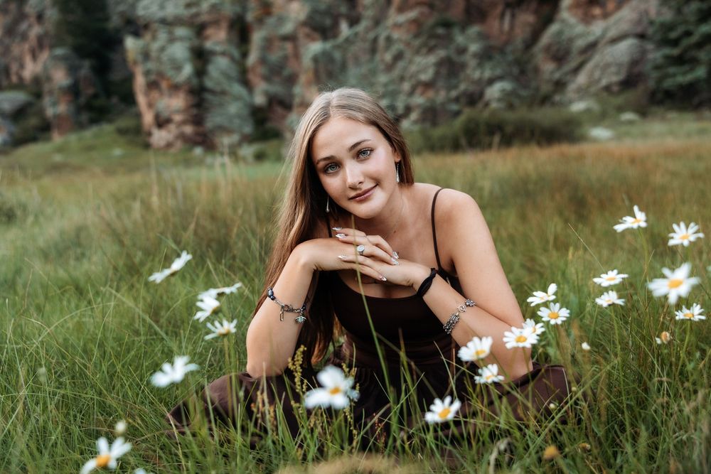 Young woman lying in a field of white daisies, smiling, wearing a dark dress with rocky cliffs behind her.