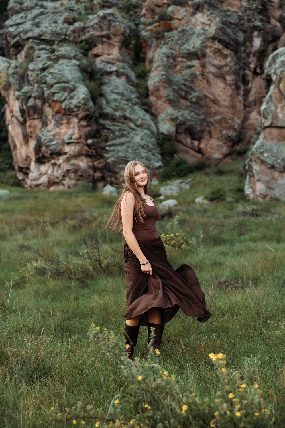 Woman in flowing brown dress walking through green meadow with dramatic rocky cliffs in background.