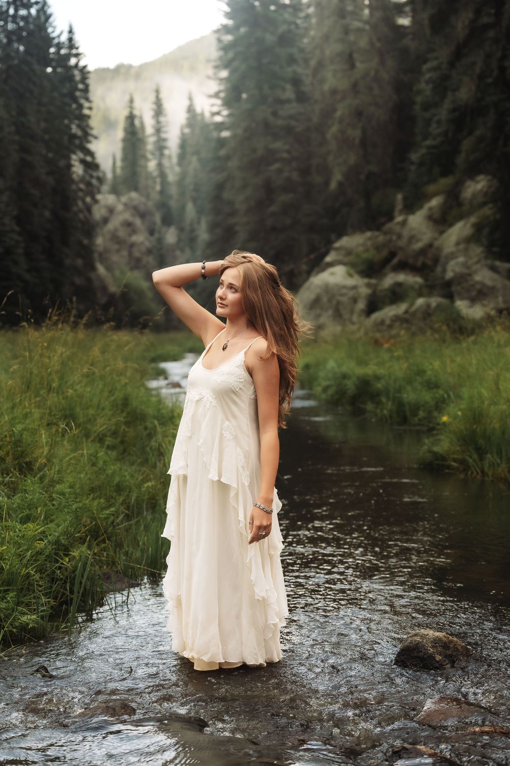 Young woman in white dress standing in a mountain stream surrounded by pine trees and rocky landscape.
