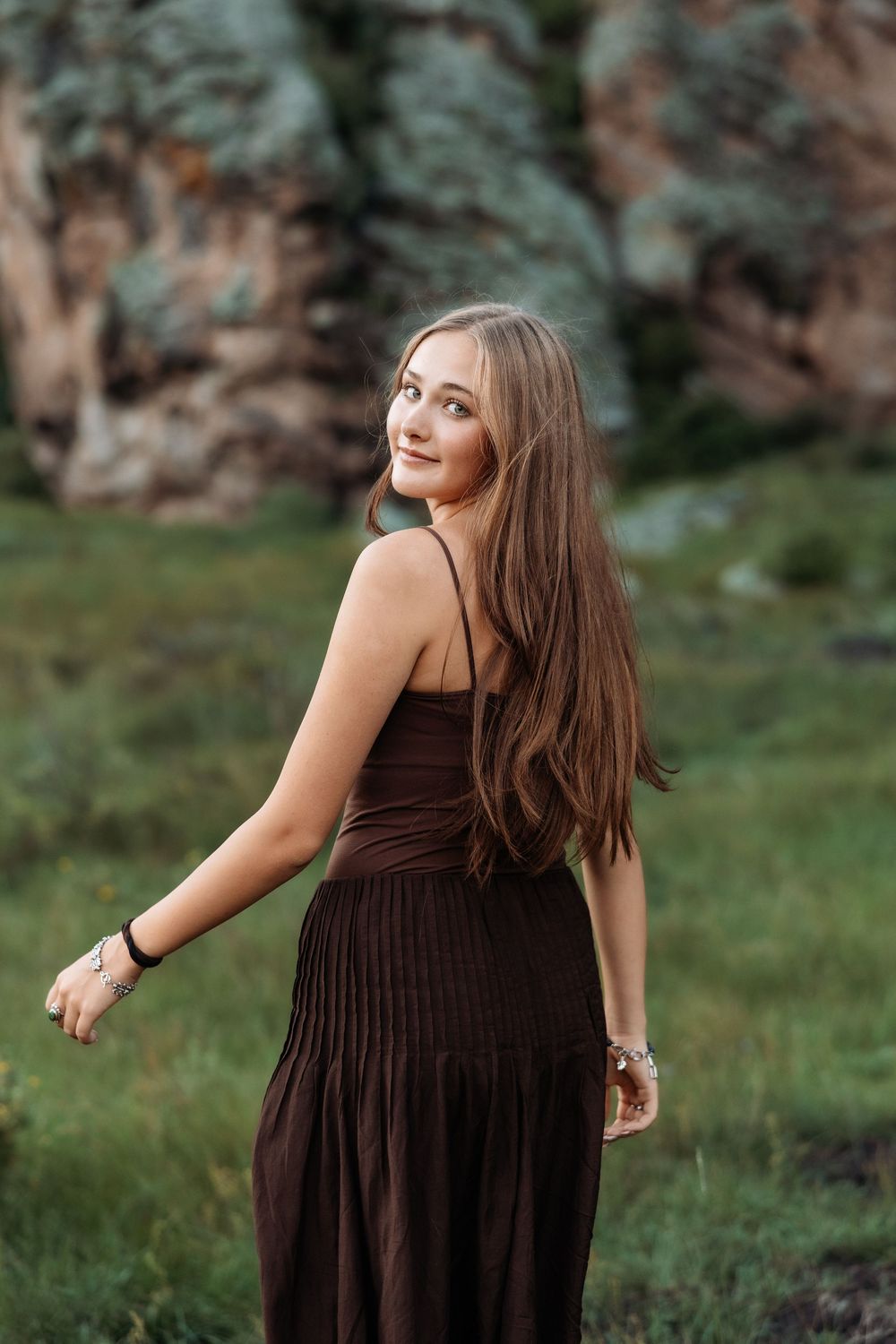 Young woman in brown dress smiling over shoulder outdoors near rocky cliff.