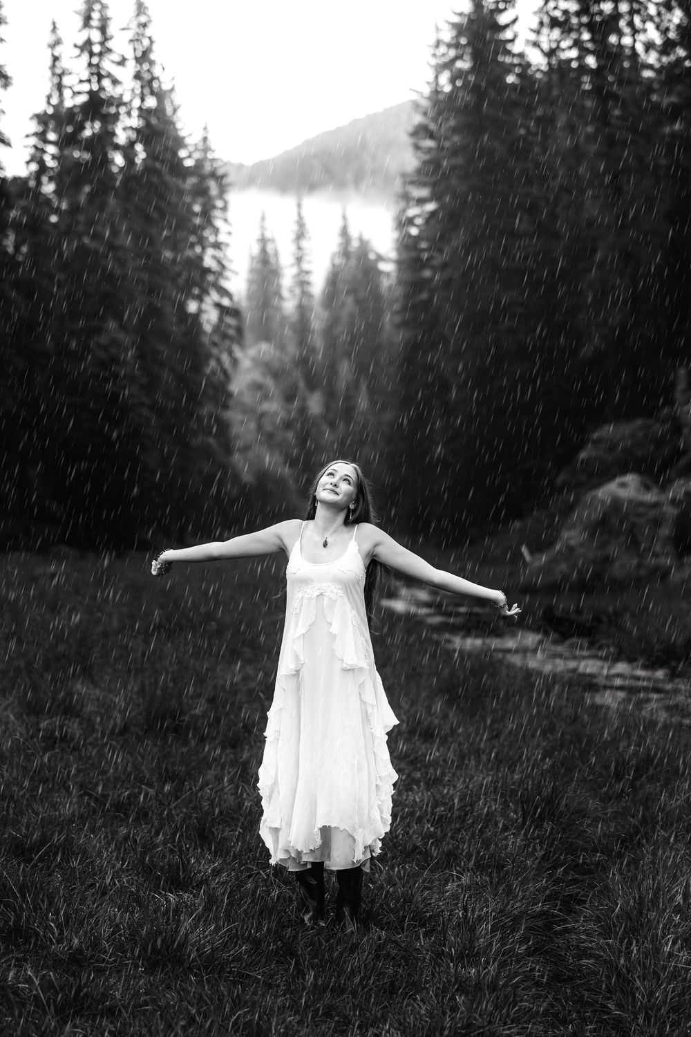 Woman in white dress embracing rain with open arms in black and white forest landscape.