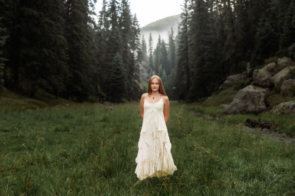 A young woman in a flowing white boho dress stands in a lush green meadow surrounded by misty pine forests.