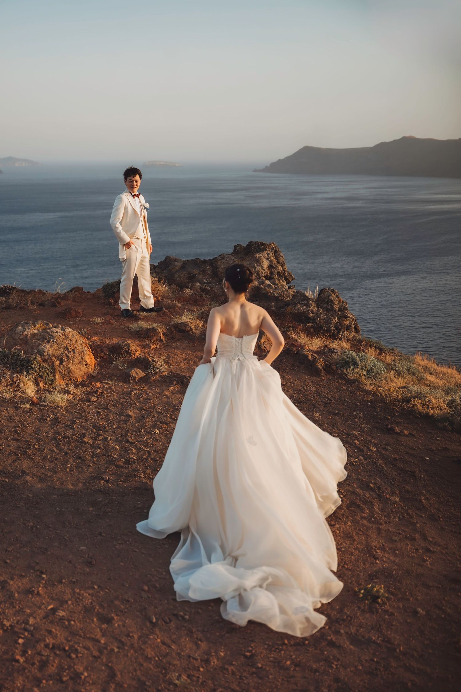Bride in flowing white gown and groom in white suit stand on cliffside overlooking ocean at sunset.