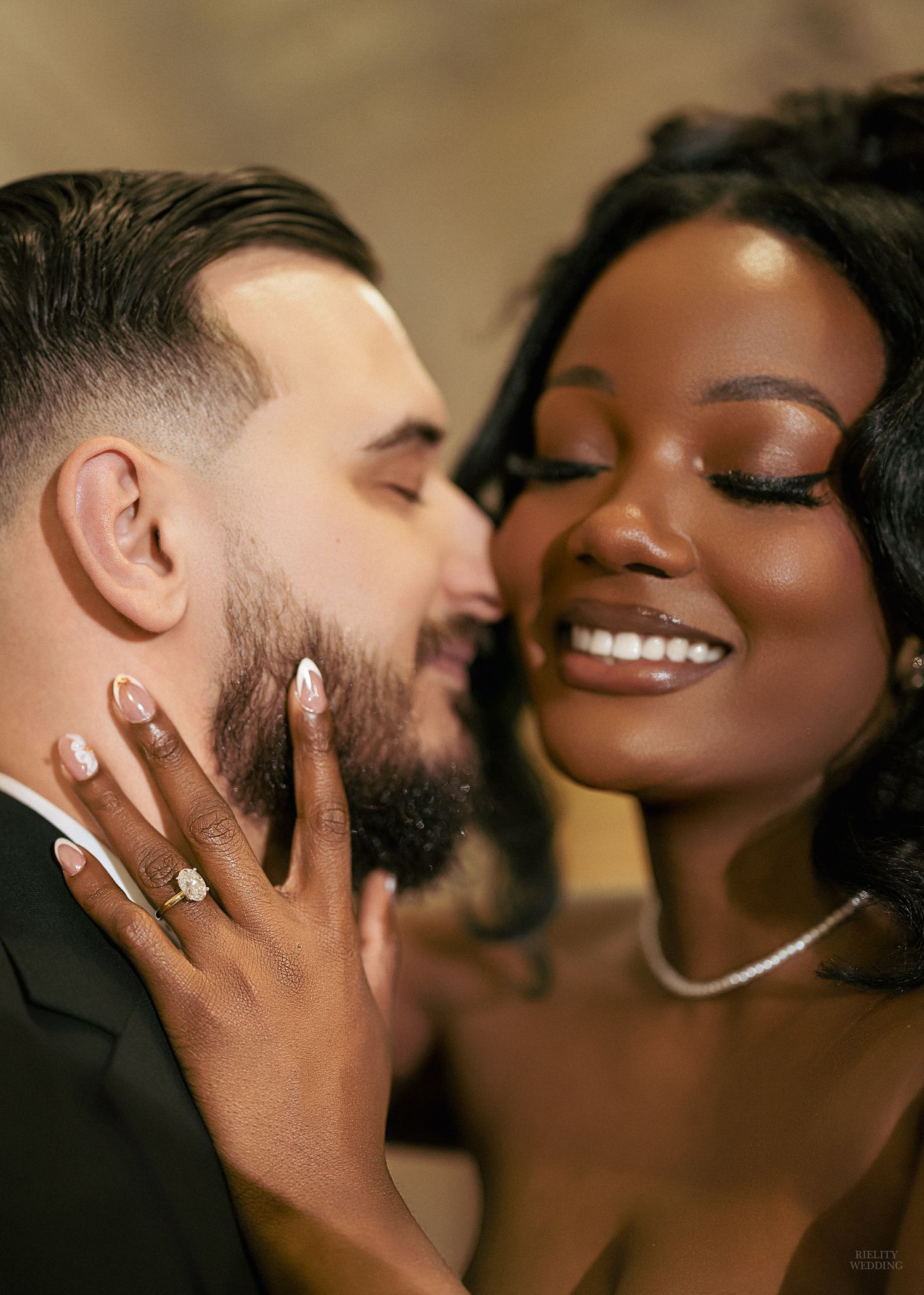 Interracial couple in wedding attire sharing a romantic moment, bride smiling with diamond ring visible.