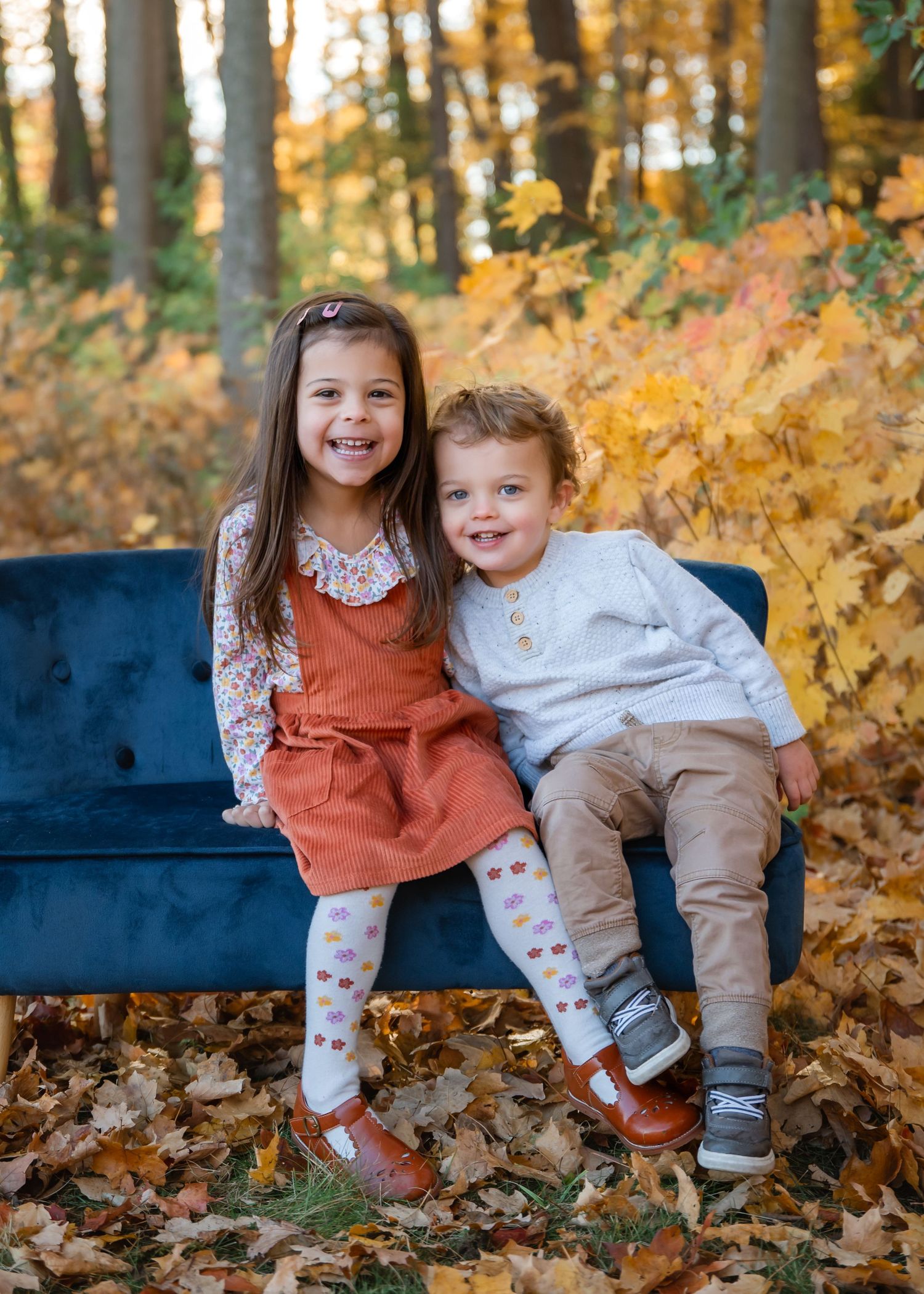Two young children smiling on a blue velvet bench surrounded by golden autumn leaves in a forest setting.
