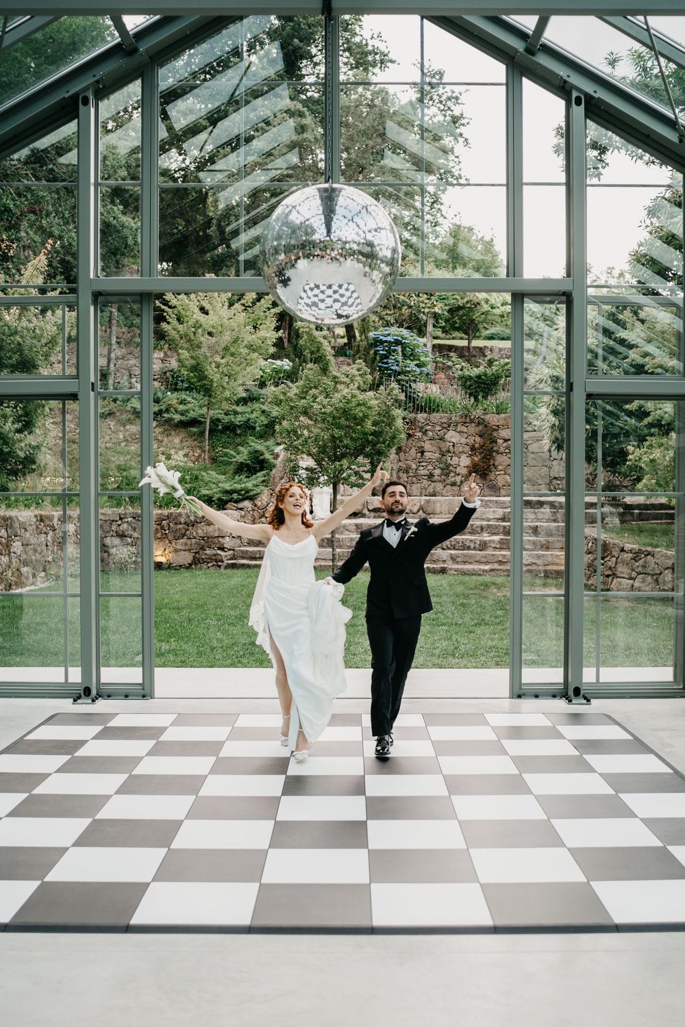 Bride and groom dancing joyfully on checkered floor in glass greenhouse venue with disco ball overhead.