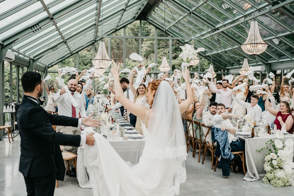 Joyful wedding reception in a glass greenhouse venue with guests raising glasses to celebrate the bride and groom.