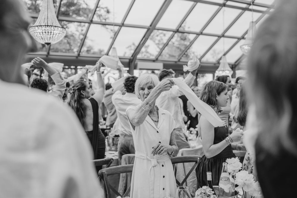 Black and white photo of wedding guests dancing and celebrating joyfully inside an elegant greenhouse venue.
