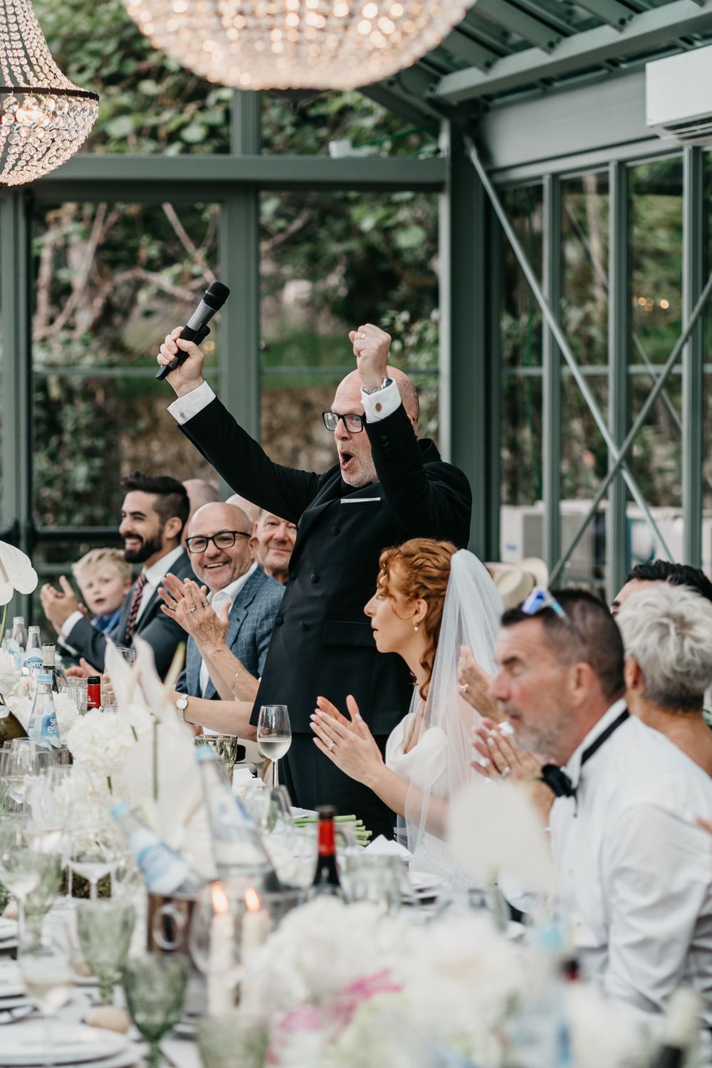 Wedding guest raises microphone triumphantly during reception toast as bride and guests applaud at elegant dinner.