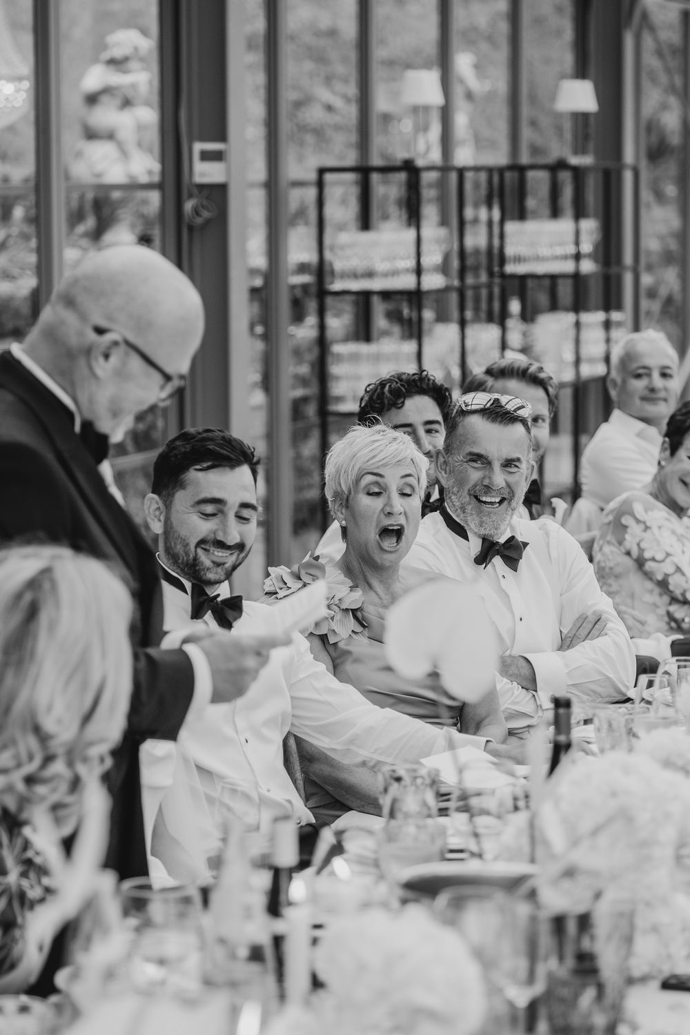 Black and white wedding reception photo of guests laughing and reacting with joy at a elegantly set dinner table.
