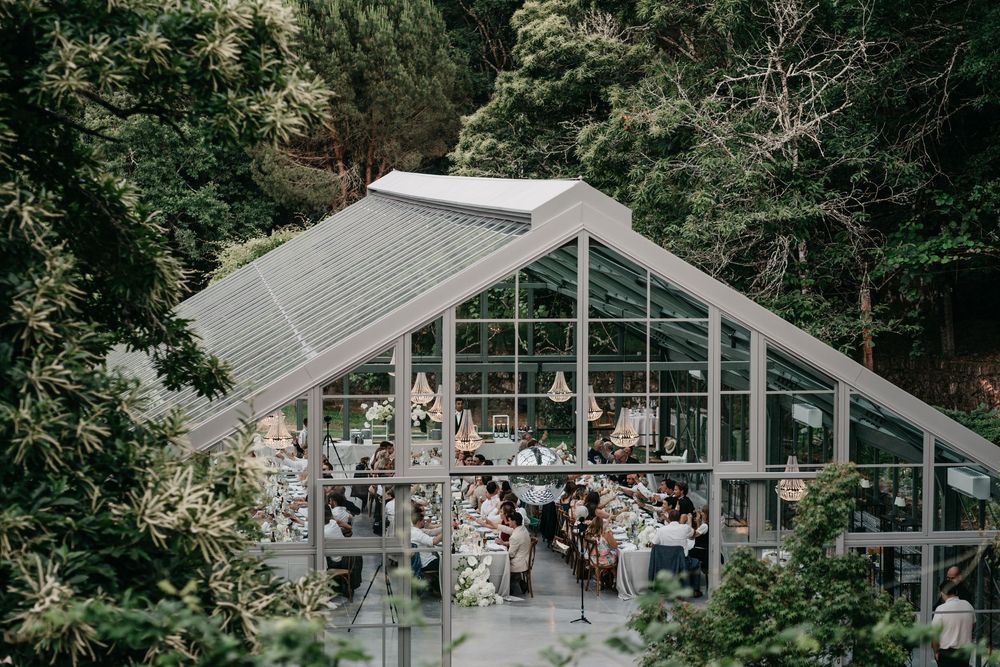 Elegant glass greenhouse wedding venue surrounded by lush green forest trees with guests dining inside.