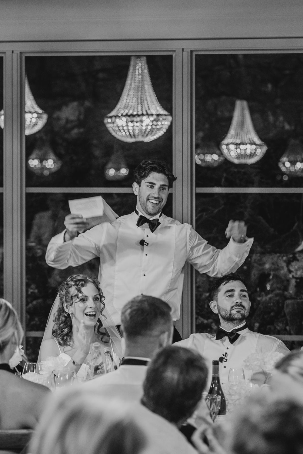 Black and white photo of a man giving a wedding speech, holding notes, with chandeliers and smiling guests in background.