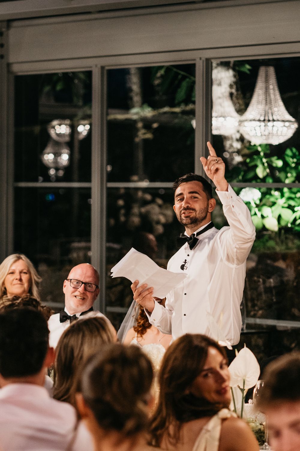 A man in a tuxedo gives a wedding speech, pointing upward, while guests smile at a elegant reception.