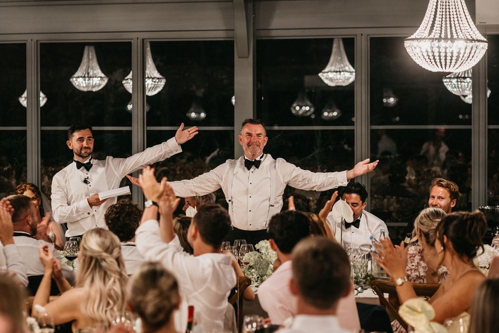 Groom celebrates with groomsmen during wedding reception toast, guests applauding under crystal chandeliers.