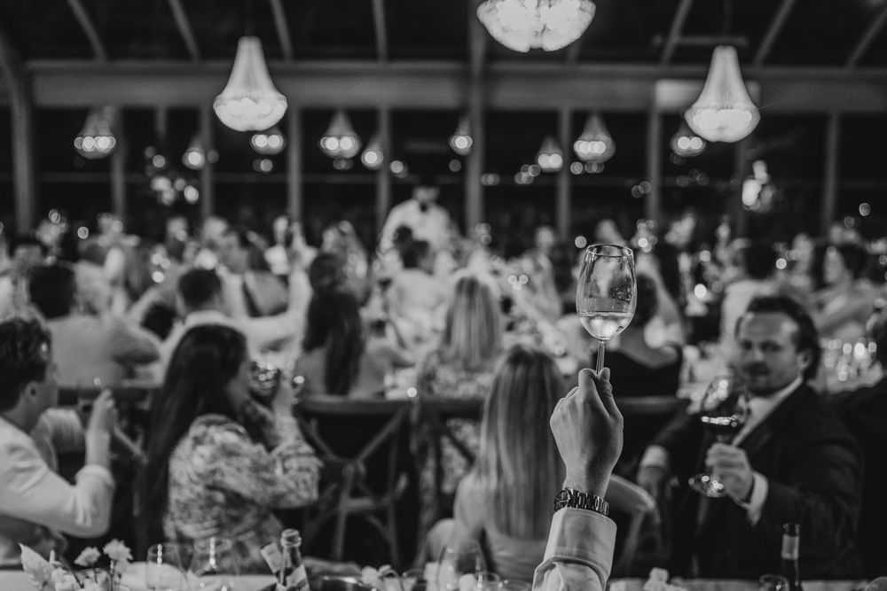 Black and white photo of guests raising wine glasses at an elegant indoor wedding reception with chandeliers.
