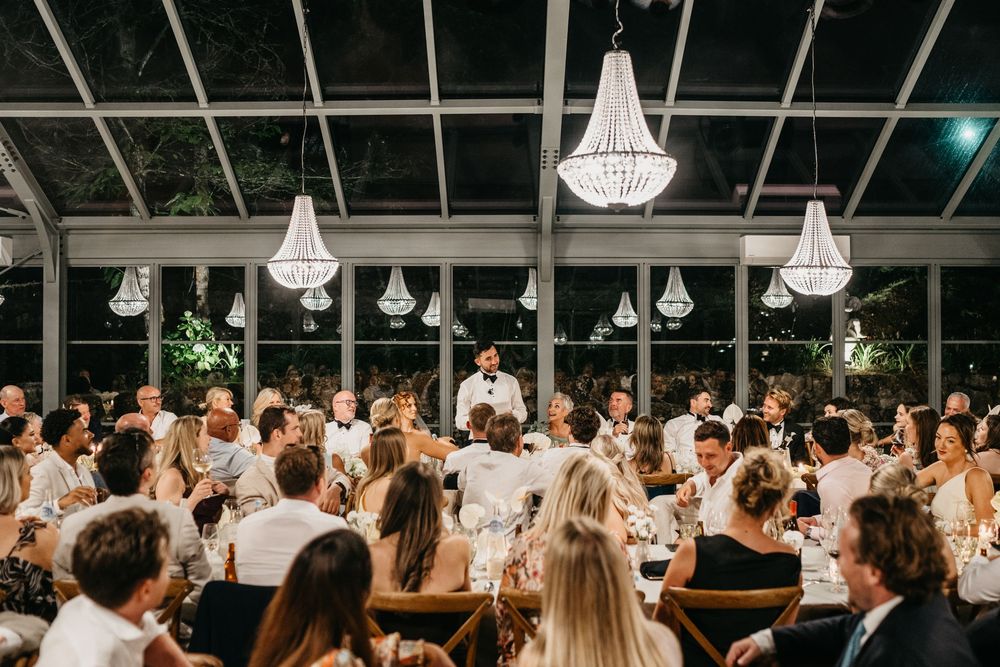 Elegant wedding reception in a glass venue with chandeliers, guests in white attire, and a speaker at the head table.