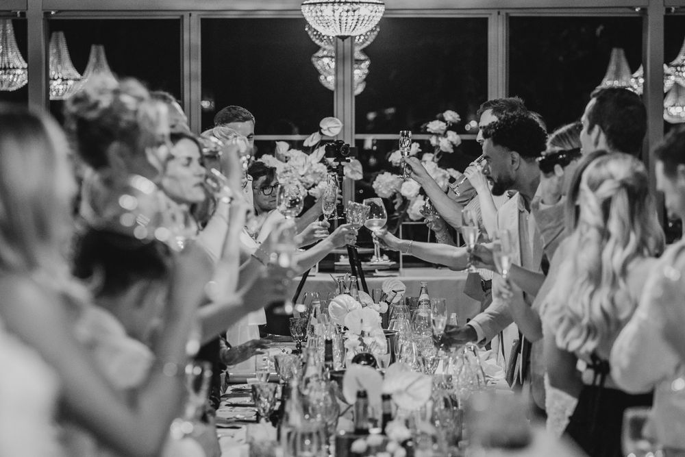 Black and white photo of wedding guests raising glasses in a toast at an elegant reception dinner.