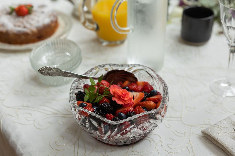 Crystal bowl filled with fresh mixed berries and fruit salad garnished with a flower, served on a white tablecloth.