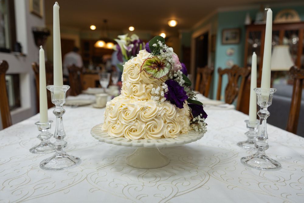 Two-tier white rosette wedding cake with purple flowers on a pedestal, flanked by crystal candlesticks on a lace tablecloth.