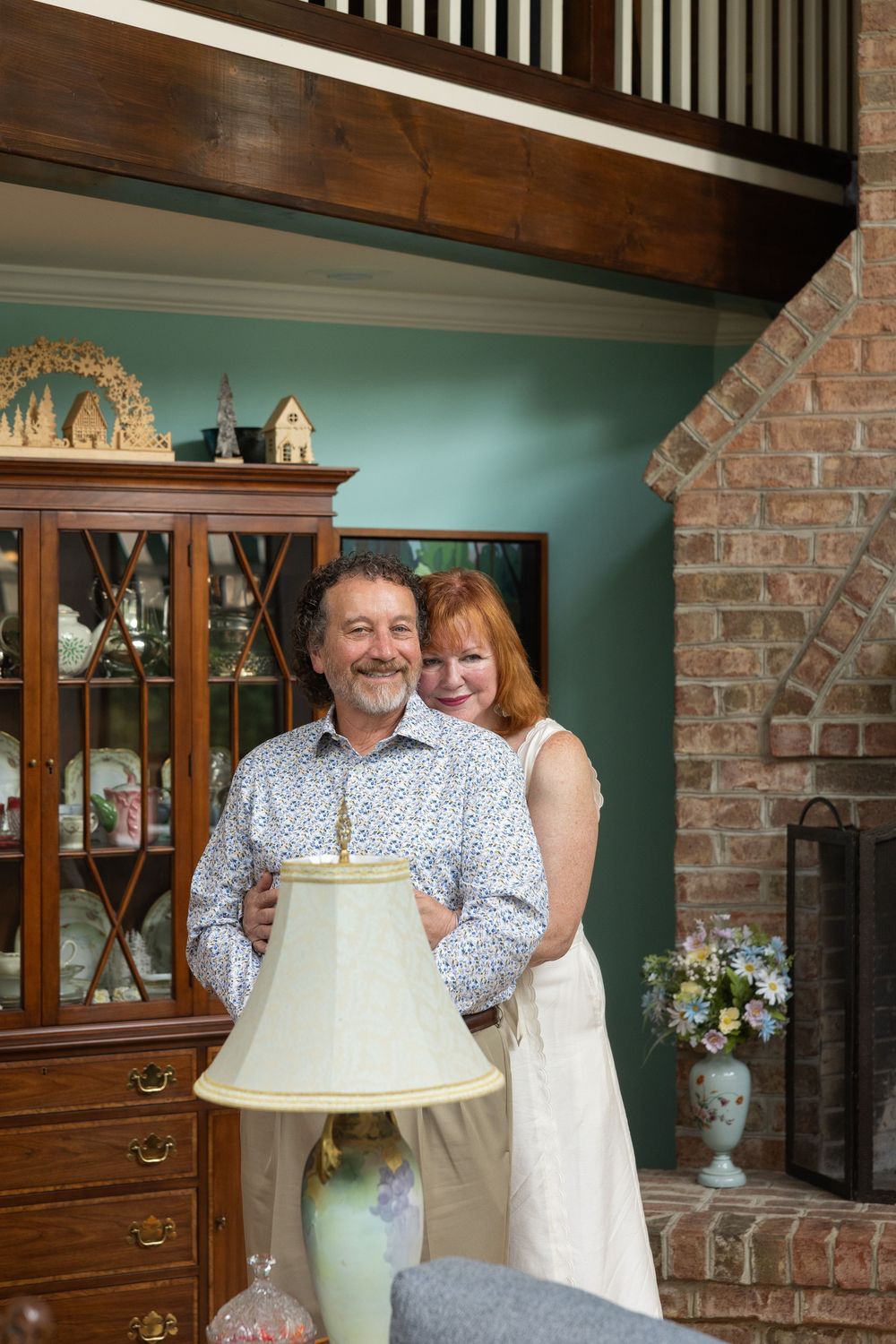 A smiling middle-aged couple poses together in a cozy living room with a brick fireplace and wooden china cabinet.