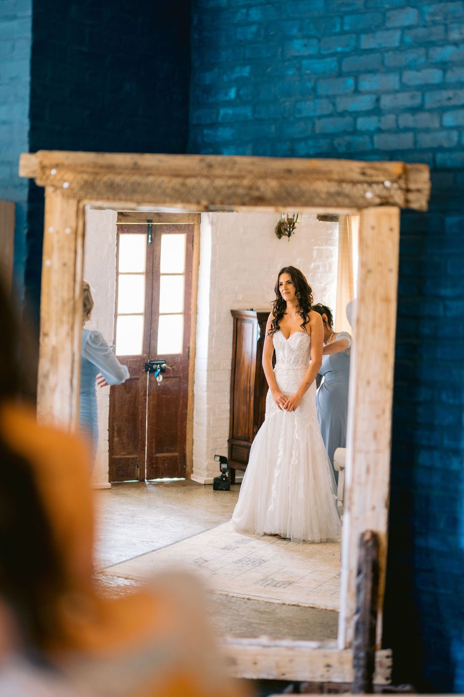 Bride in white lace wedding gown reflected in rustic wooden mirror with blue brick walls.